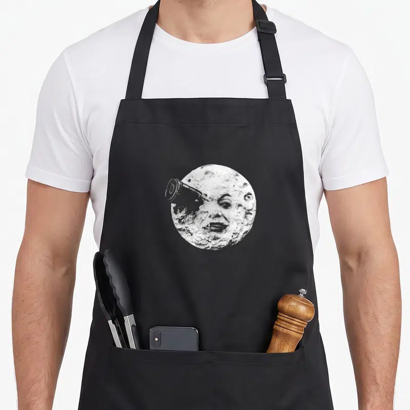 Tablier de cuisine noir avec une illustration en noir et blanc d'une lune au visage traversée par un télescope vintage.