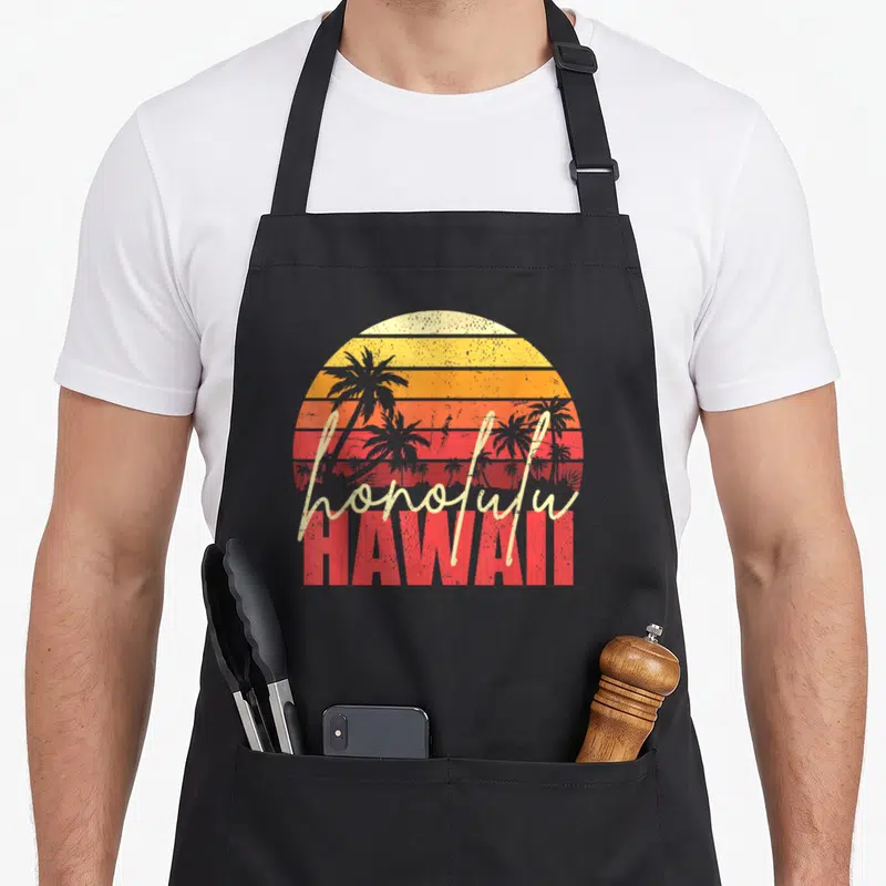 Tablier de cuisine noir avec illustration circulaire de soleil couchant, palmiers gris et texte Honolulu Hawaii en typographie vintage.