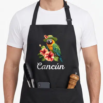 Tablier de cuisine noir orné d'un perroquet coloré coiffé d'un chapeau et de fleurs roses autour du texte blanc Cancún.