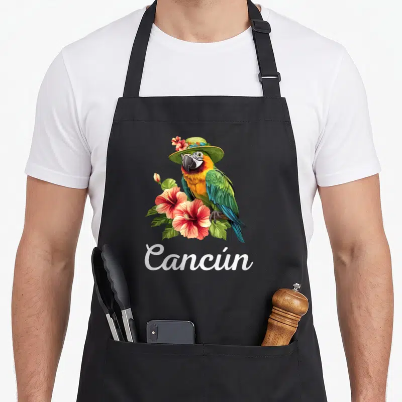 Tablier de cuisine noir orné d'un perroquet coloré coiffé d'un chapeau et de fleurs roses autour du texte blanc Cancún.