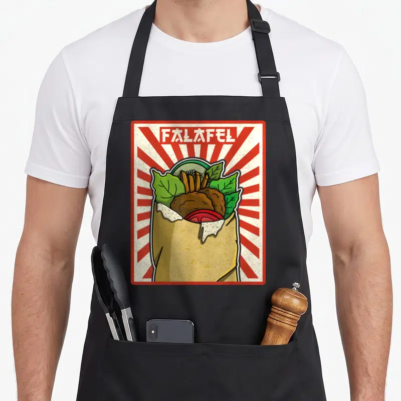Tablier de cuisine noir avec illustration colorée d'un falafel dans une pita garnie de légumes sur fond rayé rouge et blanc.