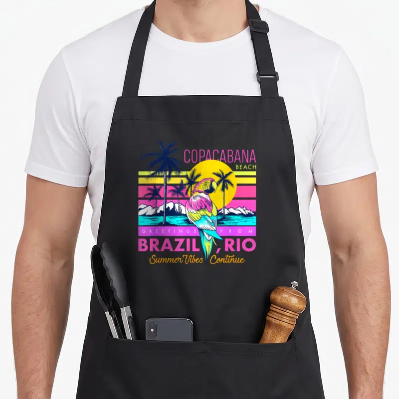 Tablier de cuisine noir avec un perroquet coloré devant un soleil jaune et des palmiers sur fond de plage tropicale.