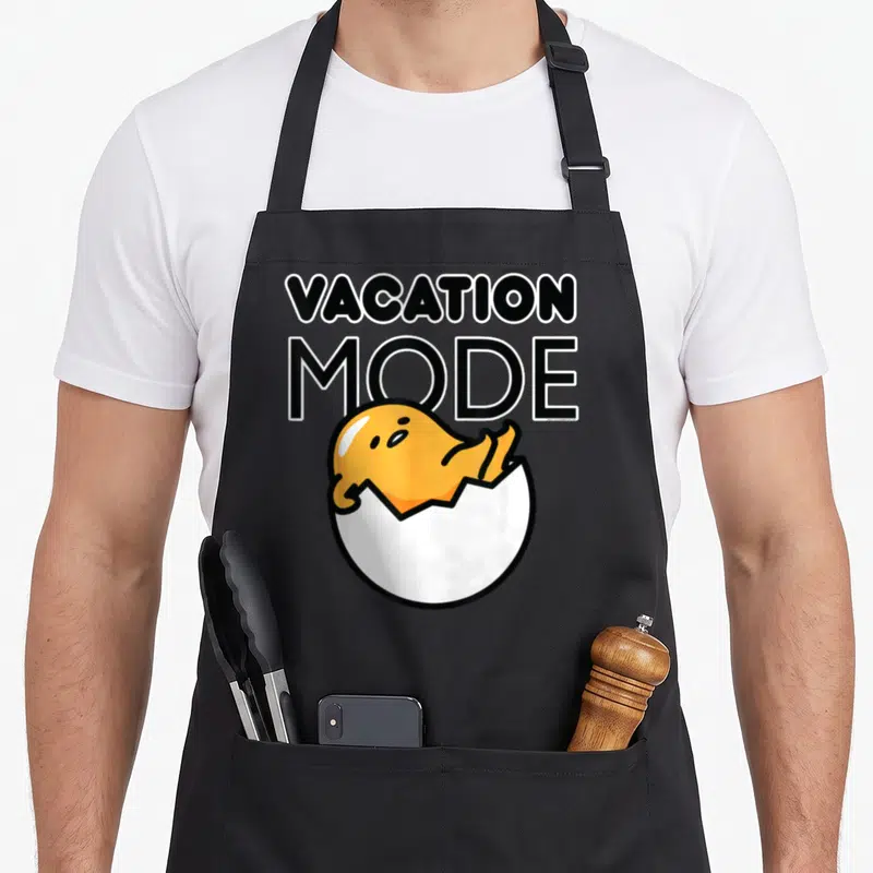 Tablier de cuisine noir avec illustration d'un œuf jaune reposant dans sa coque sous le texte Vacation Mode.