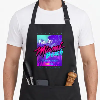 Tablier de cuisine noir avec design graphique coloré I'm in Miami Beach et palmiers stylisés en violet et turquoise.
