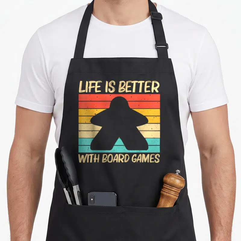 Tablier de cuisine noir avec silhouette de Meeple sur fond de bandes colorées vintage et texte Life is Better with Board Games.