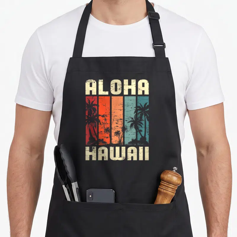 Tablier de cuisine noir avec inscription Aloha Hawaii et silhouettes de palmiers sur fond de bandes colorées vintage.