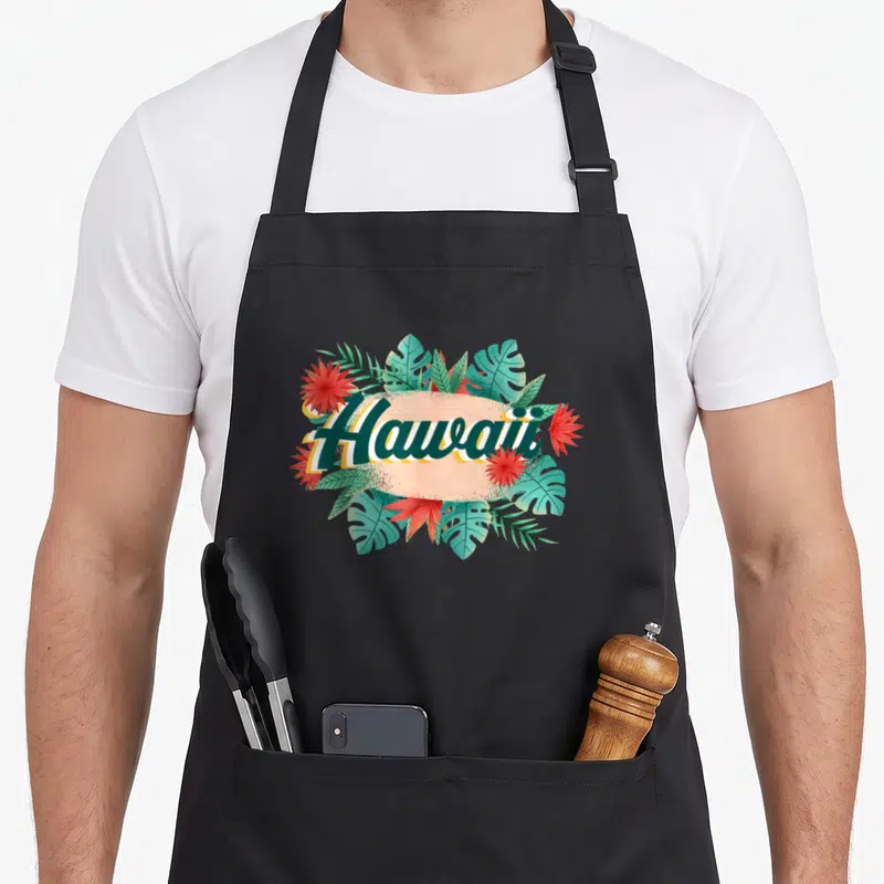 Tablier de cuisine noir avec illustration Hawaii entourée de feuilles vertes et fleurs rouges tropicales.