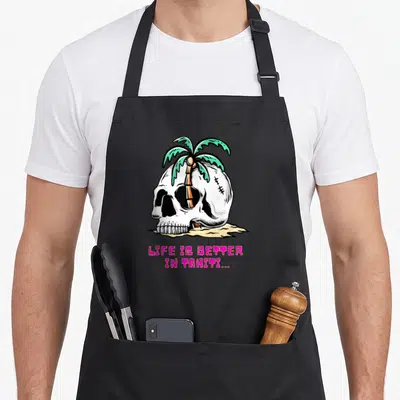 Tablier de cuisine noir avec une tête de mort blanche et un palmier vert sur fond de sable jaune.