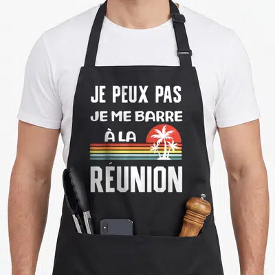 Tablier de cuisine noir avec inscription Je peux pas je me barre à la Réunion et palmiers sur fond de soleil coloré.