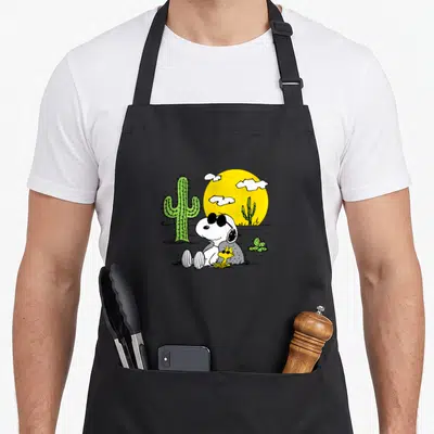 Tablier de cuisine noir avec Snoopy en lunettes noires et Woodstock assis devant un grand soleil jaune et des cactus verts.