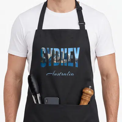 Tablier de cuisine noir avec le mot Sydney en lettrage photo contenant l'Opéra et le skyline, Australia écrit en script bleu clair en dessous.