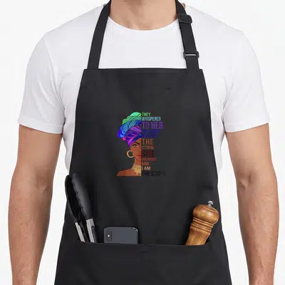 Tablier de cuisine noir avec une femme au turban multicolore et le texte They whispered to her you can't withstand the storm.