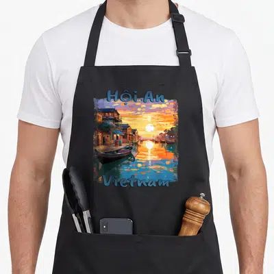Tablier de cuisine noir avec illustration colorée du coucher de soleil à Hội An au Vietnam et des bateaux traditionnels sur l'eau.