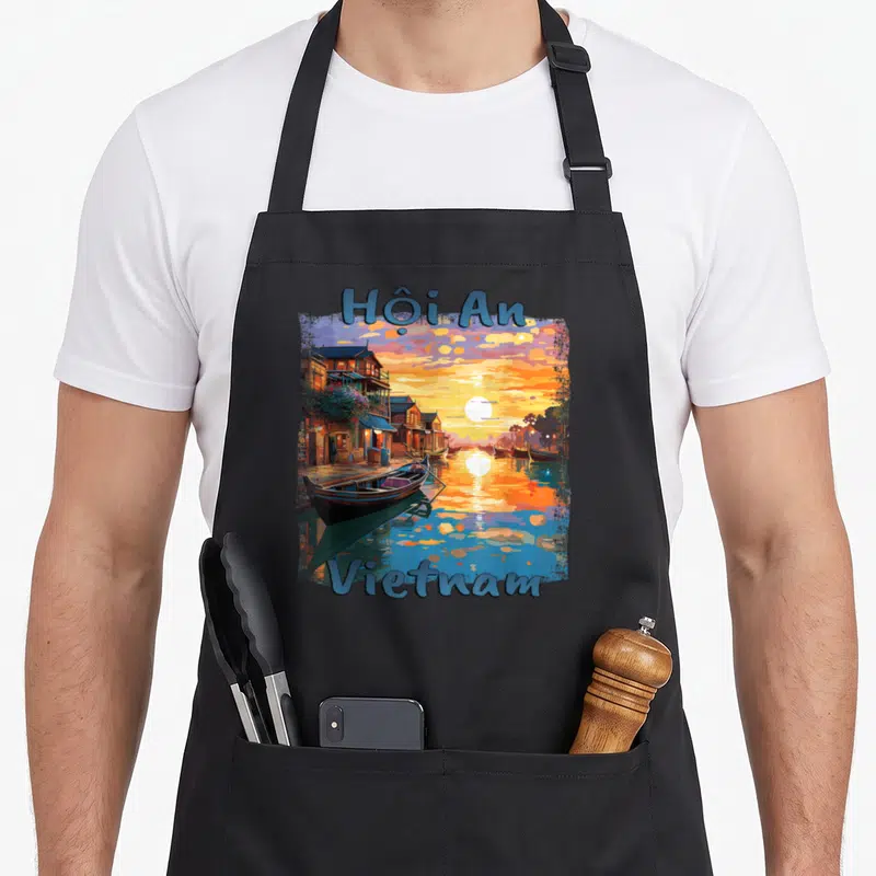 Tablier de cuisine noir avec illustration colorée du coucher de soleil à Hội An au Vietnam et des bateaux traditionnels sur l'eau.