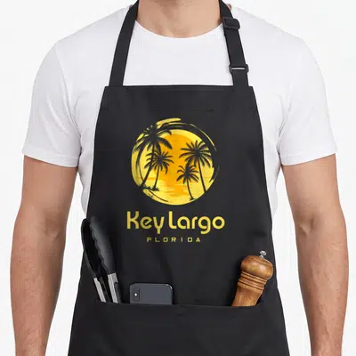 Tablier de cuisine noir avec illustration circulaire de Key Largo Florida et palmiers au coucher du soleil.