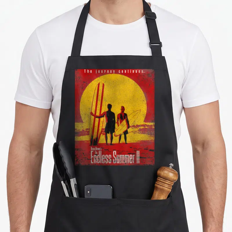 Tablier de cuisine noir avec illustration vintage de deux silhouettes de surfeurs devant un grand soleil jaune et rouge.