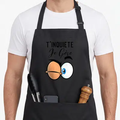 Tablier de cuisine noir avec illustration d'yeux exagérés et texte T'inquiète Je Gère en typographie noire.