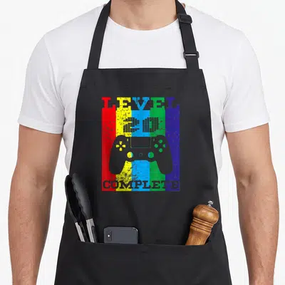 Tablier de cuisine noir avec une manette de jeu vidéo grise sur un fond à bandes colorées verticales.