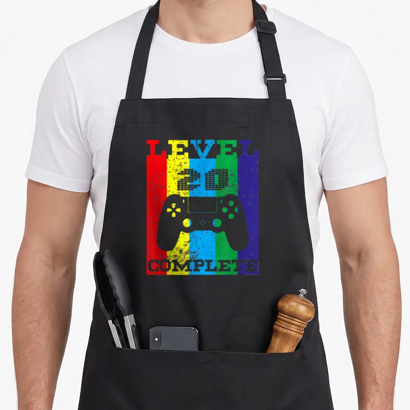 Tablier de cuisine noir avec une manette de jeu vidéo grise sur un fond à bandes colorées verticales.