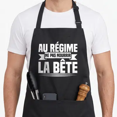 Tablier de cuisine noir avec inscription blanche Au Régime Ne Pas Nourrir La Bête sur fond uni.