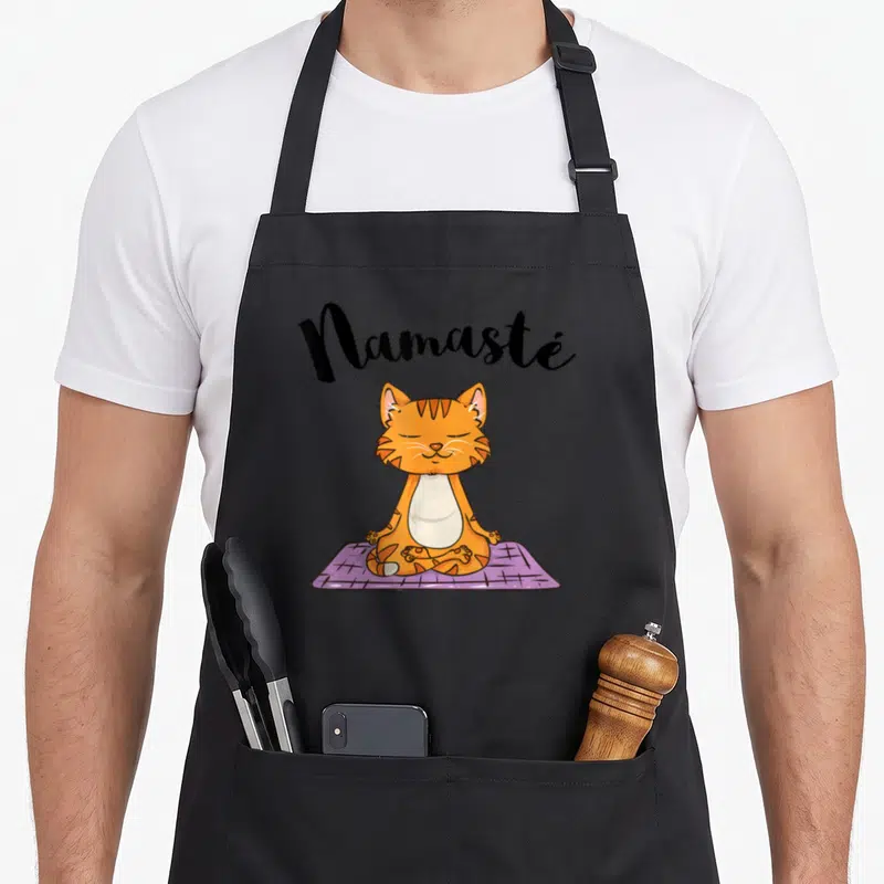 Tablier de cuisine noir avec un chat tigré orange en posture de yoga sur un tapis violet et l'inscription Namasté au-dessus.