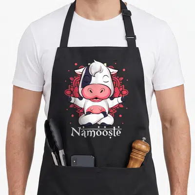 Tablier de cuisine noir avec une vache blanche et noire assise en tailleur devant un motif floral rose.