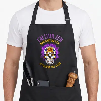 Tablier de cuisine noir avec un crâne décoré de fleurs et le texte J'ai l'air zen mais dans ma tête.