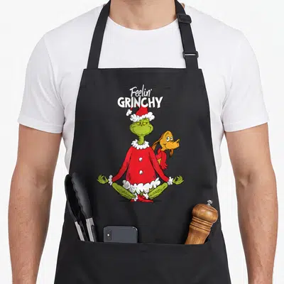 Tablier de cuisine noir avec le Grinch en tenue de yoga et Max derrière lui, texte Feelin' Grinchy en haut.