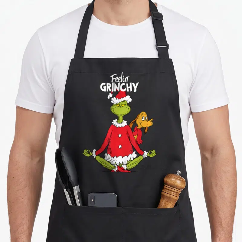 Tablier de cuisine noir avec le Grinch en tenue de yoga et Max derrière lui, texte Feelin' Grinchy en haut.