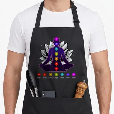 Tablier de cuisine noir avec silhouette violette en position lotus, sept chakras colorés alignés verticalement et motifs floraux blancs en arrière-plan.