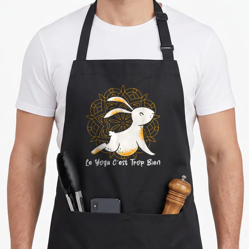Tablier de cuisine noir avec un lapin blanc en position de yoga devant un motif géométrique orange et le texte Le Yoga C'est Trop Bien.