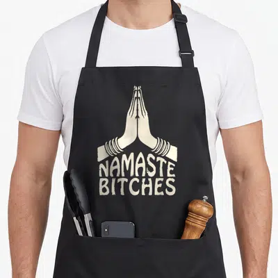 Tablier de cuisine noir avec illustration de mains jointes en prière et texte Namaste Bitches en blanc cassé.