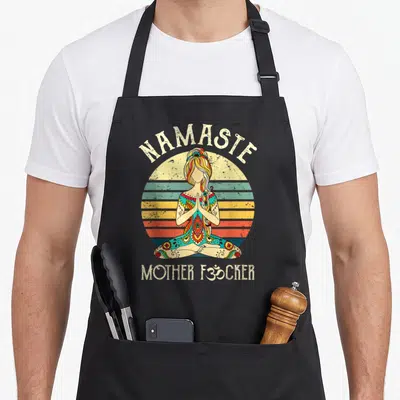 Tablier de cuisine noir avec illustration vintage d'une femme en position de yoga devant un soleil couchant coloré et des textes humoristiques.