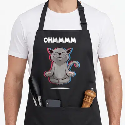 Tablier de cuisine noir avec un chat gris assis en taillet, yeux fermés, entouré de contours rose et bleu, sous le mot Ohmmm en lettres blanches.