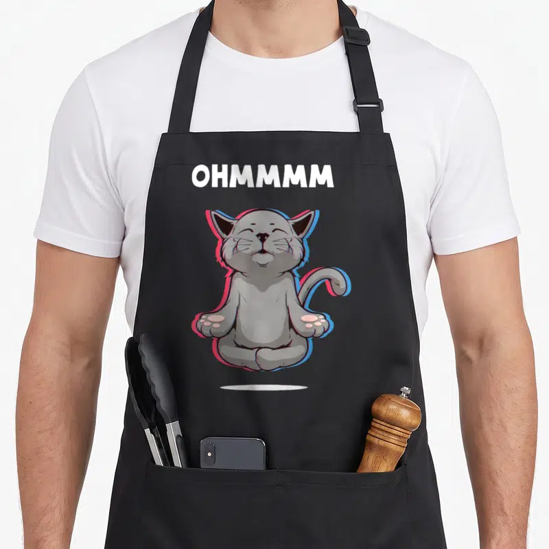 Tablier de cuisine noir avec un chat gris assis en taillet, yeux fermés, entouré de contours rose et bleu, sous le mot Ohmmm en lettres blanches.
