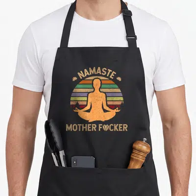 Tablier de cuisine noir avec une silhouette orange en posture de yoga devant un soleil arc-en-ciel et le texte Namaste Motherfucker en doré.
